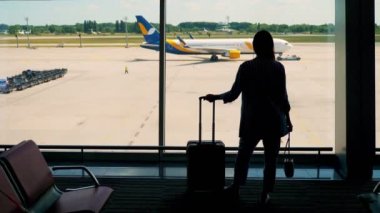 Havaalanı terminalinde panoramik pencerenin önünde duran bavullu bir kadın silueti. Açık havada büyük bir uçağa bakıyorum..