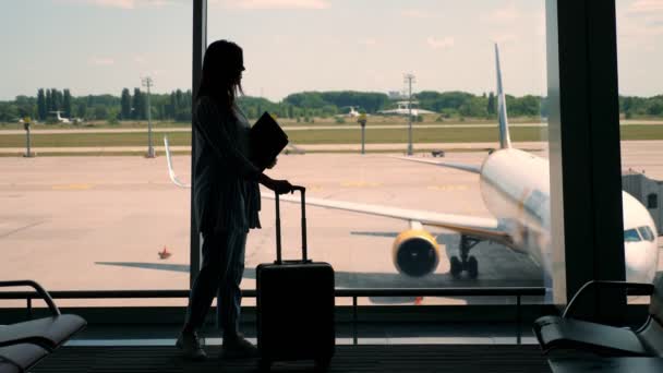 Темный силуэт деловой женщины с сумкой и ноутбуком в руках, стоящей у панорамного окна, у терминала аэропорта. глядя на большой самолет под открытым небом. — стоковое видео