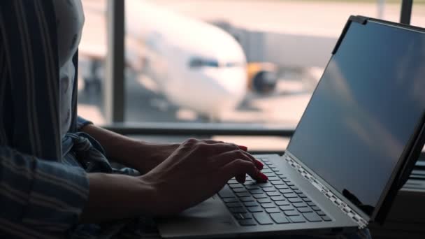 Close-up, vrouwelijke handen typen op een laptop tegen een panoramische raamachtergrond. met uitzicht op het passagiersvliegtuig, op de luchthaven. — Stockvideo