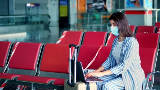 Mujer con máscara protectora, con equipaje, trabajando en la computadora portátil en el aeropuerto, mientras espera el embarque. Responsabilidad social, protección contra el virus. — Vídeo de stock