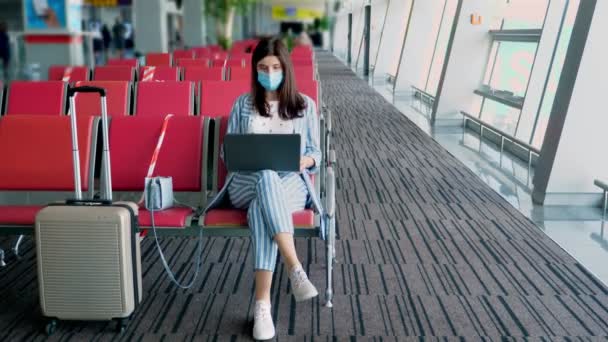 Koruyucu maskeli bir kadın, bavullu, havaalanında dizüstü bilgisayarla çalışıyor, biniş için bekliyor. Coronavirüs salgınından sonra hava yolları açılıyor. — Stok video