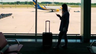 Maskeli kadın silueti, bavullu, cep telefonu kullanıyor, havaalanında panoramik pencereye karşı duruyor. Karantinadan sonra uçuşlar açıktır.