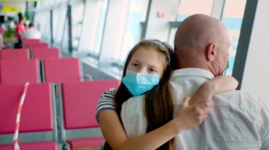 Portre, koruyucu maskeli kız babasına sarılıyor, havaalanında, kalkış salonundaki boş koltukların arkasında. Coronavirüs salgınından sonra uçuşlar başladı.