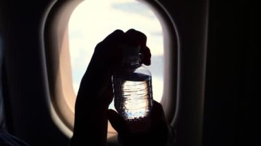 Yakın plan, uçağın penceresinin yanında elinde su şişesi tutan koyu renk siluet. Küçük plastik su şişesini bir uçak ışıklandırıcısının arka planına doğru açar..