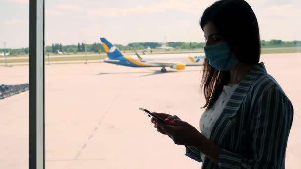 Portret van vrouw in masker, maakt gebruik van mobiel, staat tegen panoramisch raam met start-en landingsbaan en groot vliegtuig uitzicht, op de luchthaven. vluchten zijn geopend na quarantaine — Stockvideo