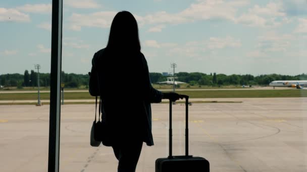 Silhouet van elegante zakenvrouw met handbagage, kijkend door het raam naar start-en landingsbaan en vertrekkend vliegtuig, op de luchthaven. — Stockvideo