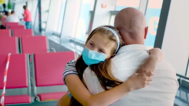 Portre, koruyucu maskeli kız babasına sarılıyor, havaalanında, kalkış salonundaki boş koltukların arkasında. Coronavirüs salgınından sonra uçuşlar başladı. — Stok video