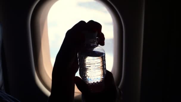 Close-up, σκούρο σιλουέτα του χέρι κρατώντας μπουκάλι νερό κοντά στο παράθυρο του αεροπλάνου. ανοίγει μικρή πλαστική φιάλη νερού, στο φόντο ενός φωτιστή αεροπλάνου. — Αρχείο Βίντεο