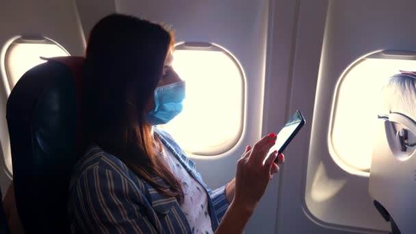 Wanita muda bertopeng pelindung menggunakan ponsel pintar di dalam pesawat, duduk di dekat penerang. Melanjutkan penerbangan setelah pandemi coronavirus. Membuka perbatasan. Perjalanan pasca karantina.. — Stok Video