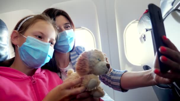 Koruyucu maskeli, çocuklu bir kadın. Akıllı telefon cihazıyla video bağlantısı kuruyor. Uçağın içinde otururken video çekmek ya da selfie çekmek. Coronavirüs salgınından sonra uçuşlar başladı.. — Stok video