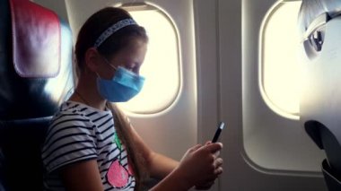 Kız, koruyucu maskeli çocuk cep telefonu kullanıyor, akıllı telefon uçağın içinde, aydınlatma cihazının yanında oturuyor. Coronavirüs salgınından sonra uçuşlar devam ediyor. Sınırlar açılıyor. Karantina sonrası seyahat.