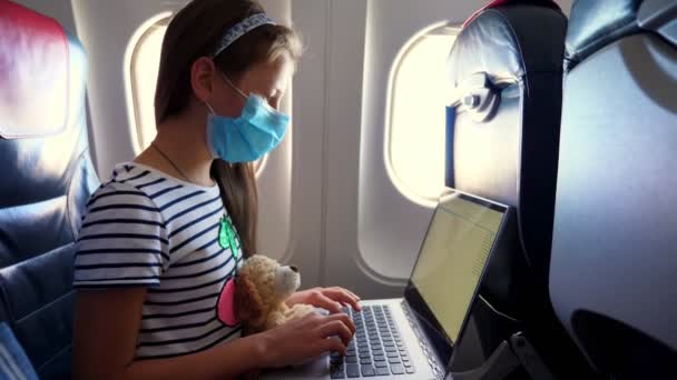 Niña, niño en máscara protectora utiliza el ordenador portátil mientras viaja en avión. sentado cerca de la ventana en el avión. vuelos abiertos después de la epidemia de coronavirus. educación a distancia, escuela en línea y concepto de viaje. — Vídeos de Stock