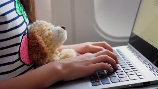 Κοντινό πλάνο των χεριών, το παιδί πληκτρολογεί σε ένα πληκτρολόγιο laptop, μέσα σε ένα αεροπλάνο, κάθεται δίπλα σε ένα φωτιστικό, κρατώντας ένα μικρό παιχνίδι. πτήσεις που άνοιξαν μετά την επιδημία του Coronavirus. εξ αποστάσεως εκπαίδευση, σχολείο — Αρχείο Βίντεο