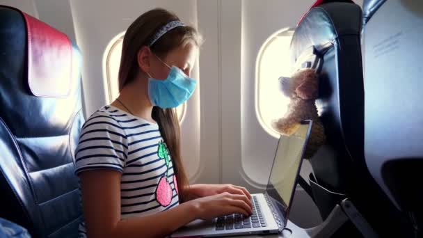 Fille, enfant dans un masque de protection utilise un ordinateur portable, à l'intérieur d'un avion, assis près d'un illuminateur. vols ouverts après l'épidémie de coronavirus. enseignement à distance, école en ligne et concept de voyage. — Video