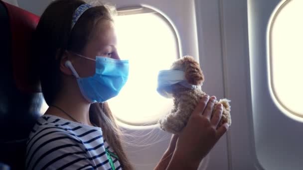 写真、保護マスクとヘッドフォンの子供の女の子は、音楽に耳を傾け、イルミネーターの近くの飛行機の中で、マスクを身に着けているおもちゃの犬と遊ぶ。コロナウイルスの後に — ストック動画