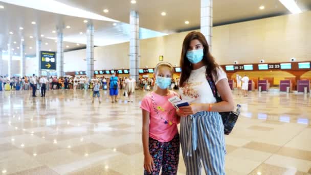 Vrouw en meisje kind met maskers op de luchthaven, tegen de achtergrond van check-in balies en vliegtuigpassagiers met bagage. vluchten die heropend zijn na de uitbraak van het coronavirus. Grenzen openen. reizen open. — Stockvideo
