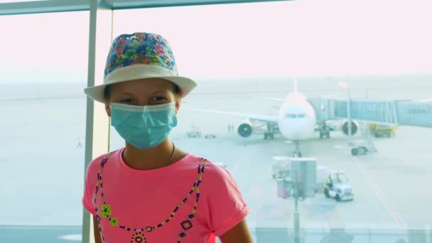 Porträtt av barn flicka i medicinsk skyddsmask på flygplatsen, framför panoramafönster med landningsbana och stora plan utsikt. flygningar som återupptas efter utbrottet av coronavirus. Öppnar gränser. resor — Stockvideo