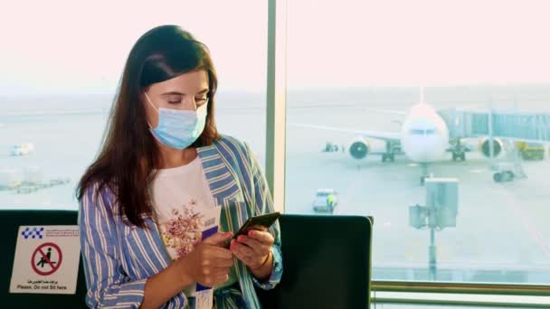 Porträtt av kvinna i mask, använder mobil, sitter framför panoramafönster med landningsbana och stora plan utsikt, på flygplatsen. flygningar som återupptas efter utbrottet av coronavirus. Öppnar gränser. resor öppna — Stockvideo
