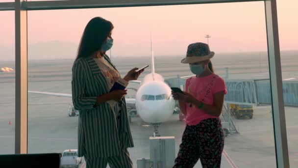 마스크를 쓴 여성과 10 대 소녀는 여권과 티켓을 소지 한 채 공항에서 파노라마 창 앞에서 휴대 전화를 사용 한다. 코로나 바이러스가 창궐 한 후 재개 된 비행 — 비디오