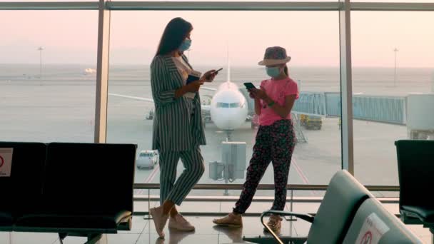 Женщина и девочка-подросток, в масках, с паспортами и билетами, пользуются мобильниками, перед панорамным окном с взлетно-посадочной полосой и видом на большой самолет, в аэропорту. возобновление рейсов после окончания вспышки коронавируса — стоковое видео