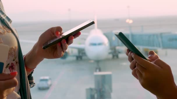 Zbliżenie rąk. kobieta i nastolatka, korzysta z telefonów komórkowych, przed panoramicznym oknem z widokiem na pas startowy i duży samolot, na lotnisku. loty wznowione po zakończeniu epidemii koronawirusu. Otwarcie granic — Wideo stockowe