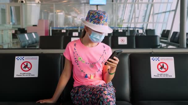 Девочка-подросток в защитной маске пользуется мобильным телефоном. Она сидела в пустом зале ожидания в аэропорту, ожидая посадки на самолет. рейсы вновь открылись после окончания вспышки коронавируса. Открываю границы. Путешествие открыто. — стоковое видео