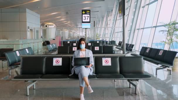 보호용 마스크를 쓰고 빈 공항 라운지에 앉아 비행기를 기다리고 있는 여성이 노트북을 사용 한다. 코로나 바이러스가 창궐 한 후에 비행기 여행이 재개되었습니다. 국경을 개방 합니다. 여행을 열다. — 비디오