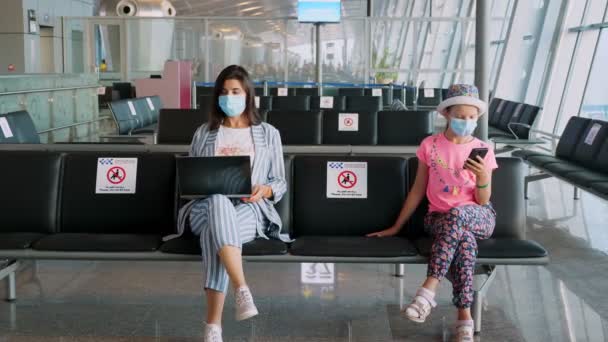 Жінка і дівчина-підліток, в захисних масках, використовують мобільний і ноутбук, сидячи в порожньому залі аеропорту, чекаючи на політ. польоти знову відкриті після закінчення коронавірусу. Відкриття кордонів — стокове відео