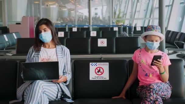 Woman and teenage girl, in protective masks, use mobile and laptop, while sitting in empty airport lounge, waiting to board a flight. flights re-opened after coronavirus outbreak end. Opening borders — Stock Video