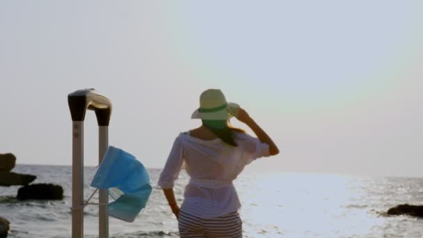 Ung kvinna i sommarkläder och solhatt, möter gryningen på stranden vid havet. Närbild av hennes bagage med medicinsk mask. sjöresa efter utbrottet av coronavirus. — Stockvideo