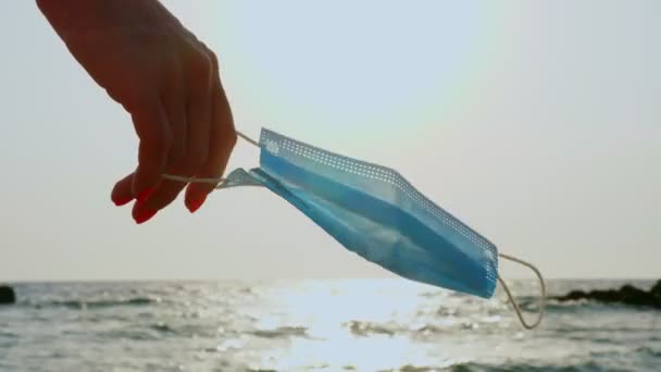 Close-up, mão feminina segura máscara médica balbuciando no vento, contra o fundo do mar. nascer do sol na praia. conceito de liberdade do coronavírus. viajar aberto — Vídeo de Stock