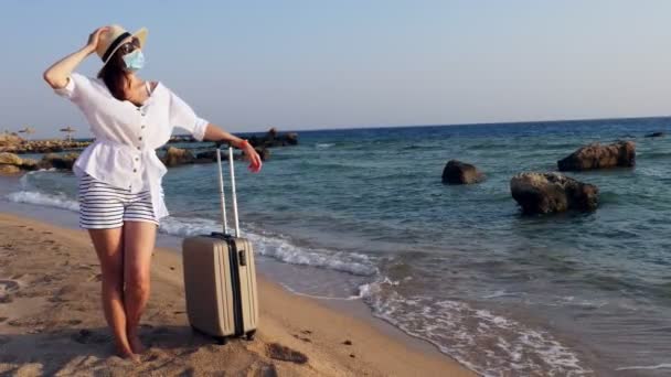 保護マスク、夏服、サングラス、サンハットの女性は、暖かい日差しを楽しみ、海のビーチで、彼女の旅行のスーツケースにもたれています。コヴィト19世の後の旅行. — ストック動画