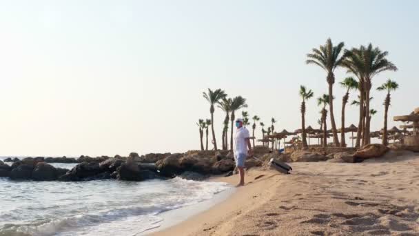 Hombre en máscara protectora y ropa de verano, disfruta de cálidos rayos de sol, junto al mar, playa con palmeras, con su maleta de viaje. el final del autoaislamiento de Covid-19. Abrir fronteras. — Vídeo de stock