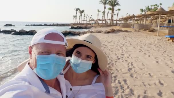 Joven hermosa pareja casada en máscaras protectoras, felizmente mirar en el marco, agitando las manos contra el fondo del mar, playa con palmeras. día cálido y soleado. concepto de vacaciones de mar con nuevas reglas — Vídeos de Stock