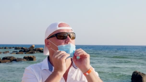 Mężczyzna w masce ochronnej i okularach przeciwsłonecznych, zdejmuje maskę i oddycha świeżym powietrzem, na tle morza, plaża z palmami. słoneczny ciepły dzień. wakacje po Covid19 samoizolacja . — Wideo stockowe