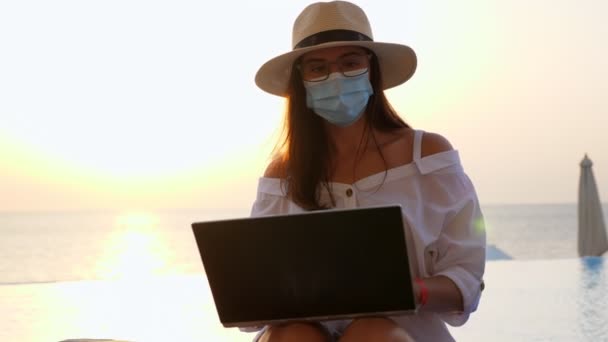 Jonge vrouw met een beschermend masker, zonnehoed en zomerkleding, werkend op laptop, zittend bij het zwembad. werken op afstand. Nieuw normaal. Leven na Covid19. — Stockvideo