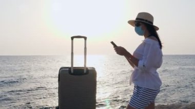 Koruyucu maskeli bir kadın, yaz kıyafetleri, güneş gözlükleri, güneş şapkası, seyahat çantası, bavulu, deniz kıyısında cep telefonu kullanıyor. Covid-19 'un peşinden gidiyorum. kendi kendini izole etmenin sonu .