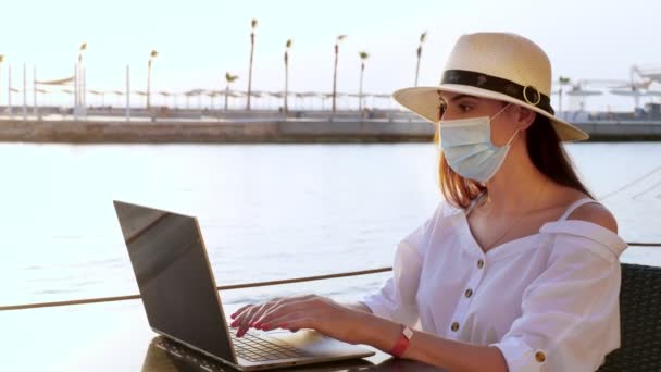 Mujer joven con máscara protectora, sombrero de sol y ropa de verano, trabajando en el ordenador portátil, sentado en un terraplén vacío junto al mar. trabajar remotamente. Viajar después de Covid-19. vacaciones de verano. — Vídeo de stock