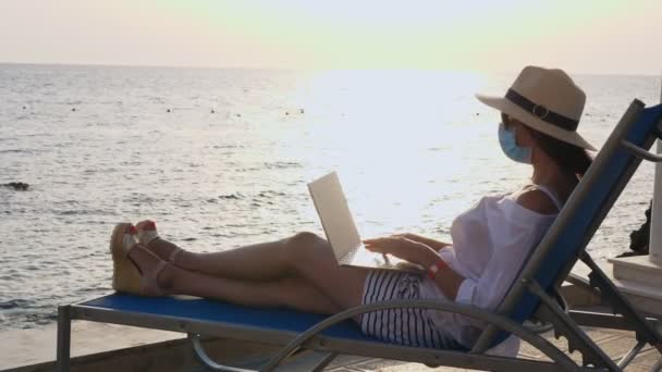 Mujer joven en máscara protectora, sombrero de sol y ropa de verano, trabajando en el ordenador portátil, sentado en la tumbona junto al mar. trabajar remotamente. nueva normalidad. Vida después de Covid19. — Vídeos de Stock