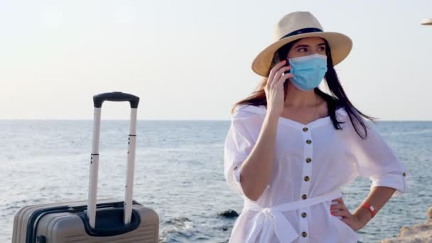 Жінка в захисній масці, літньому одязі, сонцезахисних окулярах, сонцезахисному капелюсі, з валізою для подорожей, багажем, розмовляючи на мобільному, біля берега. Поїздка за Ковидом-19. кінець самоізоляції. . — стокове відео