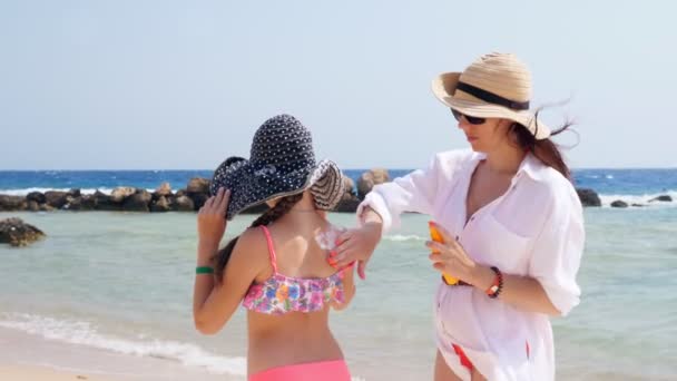 Mãe mancha sua filha, menina de 10 anos, com protetor solar, na praia sob sol quente, contra o fundo do mar. Proteção solar e UV. protecção da pele — Vídeo de Stock