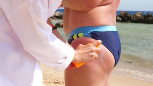 Close-up, aanbrengen van zonnebrandcrème of spraylotion op de huid van de mens. op het strand, onder hete zon, tegen de achtergrond van de zee. Zon- en UV-bescherming. huidbescherming — Stockvideo