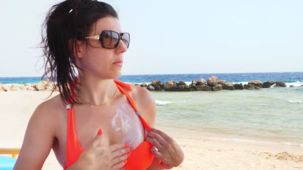 Mooie jonge vrouw, in een zonnebril, seksueel uitstrijkt, brengt zonnebrandcrème aan op de huid te ontleden. op het strand onder hete zon, tegen de achtergrond van de zee. Zon- en UV-bescherming. huidbescherming — Stockvideo