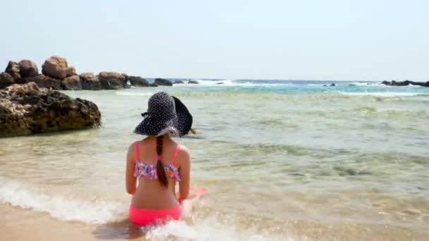 Çocuk kız, sırtı kameraya dönük, kumlu bir sahilde, denizin sıcacık suyunun tadını çıkarırken yalnız başına oturur. Yaz tatili konsepti — Stok video