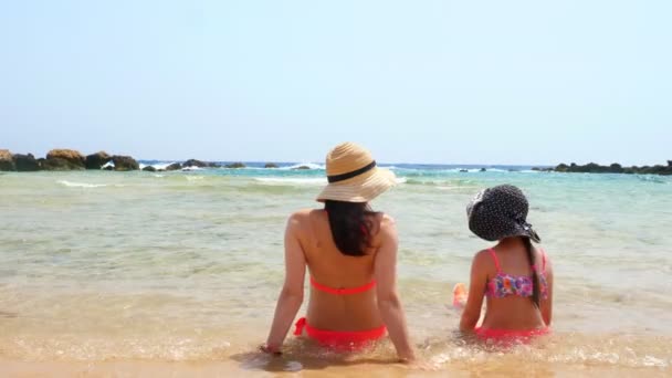 Mujer joven y niña se sientan con la espalda a la cámara, en una playa de arena, charlando, disfrutando del agua tibia del mar. verano mar vacaciones concepto — Vídeos de Stock