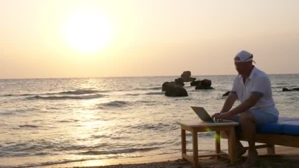 Man met blazer hoed, typend met laptop, zittend op het strand aan zee, bij zonsondergang of zonsopgang. freelancer, werk op afstand. zomervakantie op zee. internet wist afstanden uit — Stockvideo