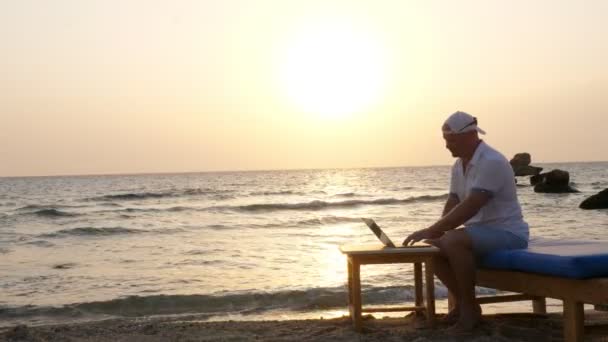 Homem de chapéu blazer, digitando usando laptop, sentado na praia junto ao mar, ao pôr do sol ou ao nascer do sol. freelancer, trabalho remoto. férias de verão no mar. internet apaga distâncias — Vídeo de Stock