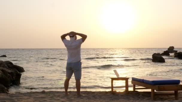 Hombre en blazer hat, disfruta del atardecer o amanecer en la playa junto al mar. un portátil abierto se encuentra junto a la tumbona. freelancer, trabajo remoto. vacaciones de verano mar. libertad con internet — Vídeos de Stock