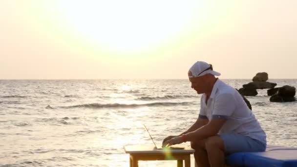Людина в капелюсі Блер, друкуючи за допомогою ноутбука, сидить на пляжі біля моря, на заході сонця або сході сонця. фрілансер, віддалена робота. літні канікули на морі. Інтернет стирає відстані — стокове відео