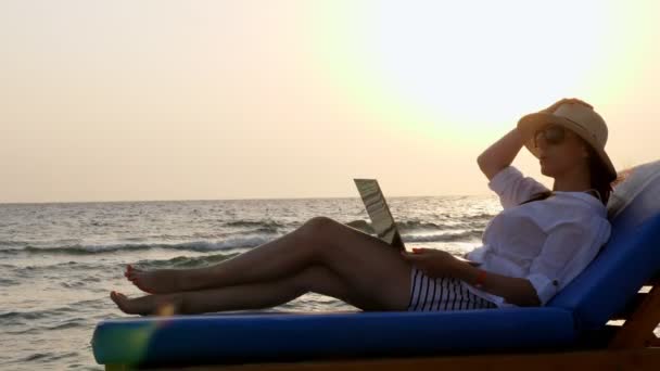 Молода жінка в сонцезахисних окулярах і сонячному капелюсі, використовує ноутбук, лежить на шезлонгу, на пляжі біля моря, на заході сонця або сході сонця. фрілансер, віддалена робота. літні канікули на морі — стокове відео